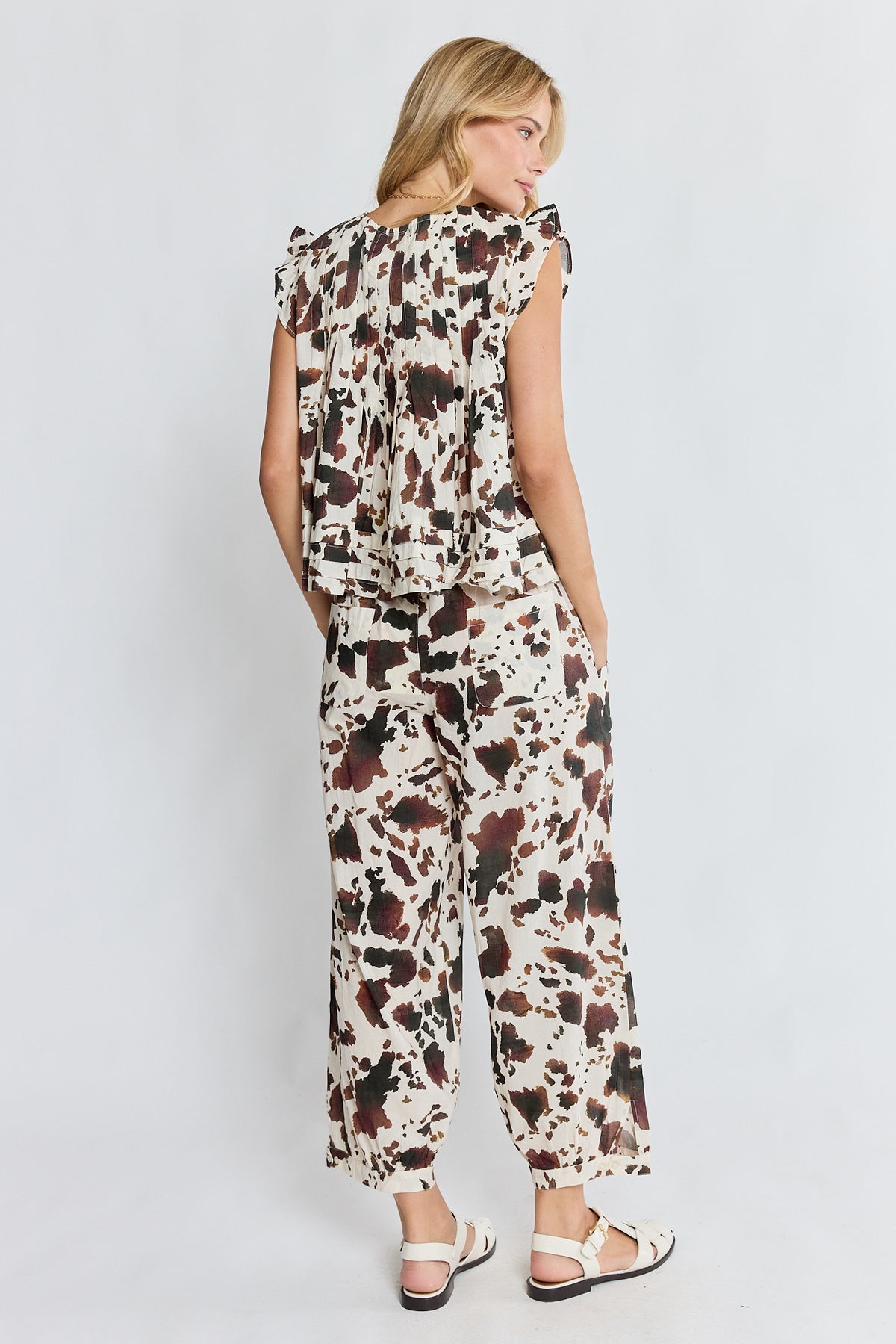 A woman shown from the back wearing a matching sleeveless ruffled top and wide-leg pants set in a brown, black, and beige cow print, paired with white sandals, against a light gray studio background.