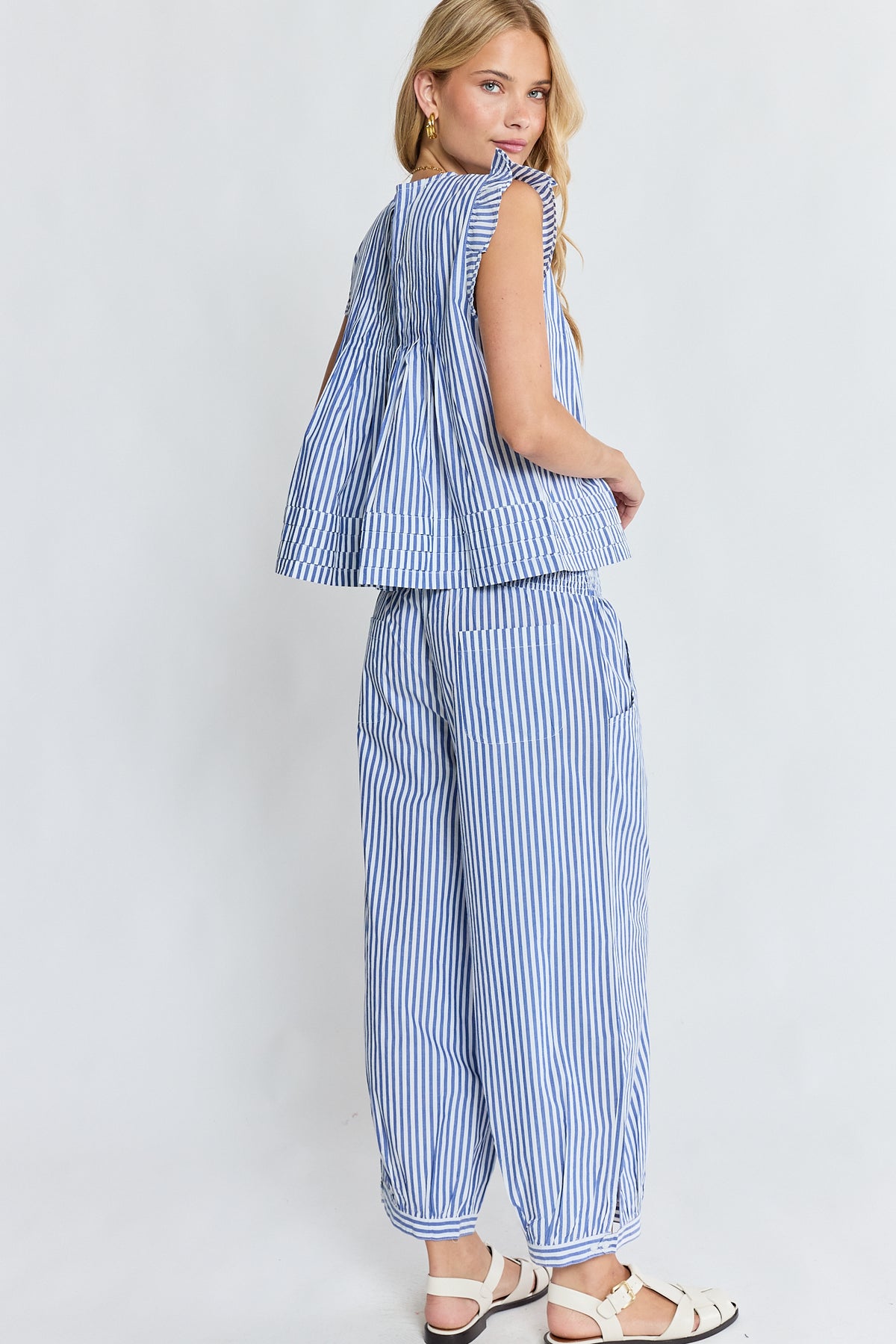 A woman modeling a blue and white striped two-piece set consisting of a sleeveless ruffled peplum top and matching wide-leg pants, paired with white sandals against a light gray studio background.