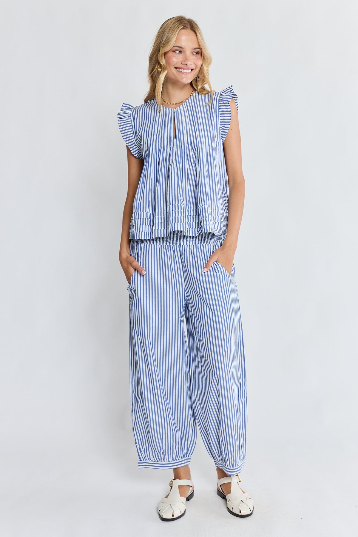 A woman modeling a matching blue and white striped two-piece set consisting of a ruffled sleeveless top and wide-leg pants, paired with white sandals, against a light gray studio background.