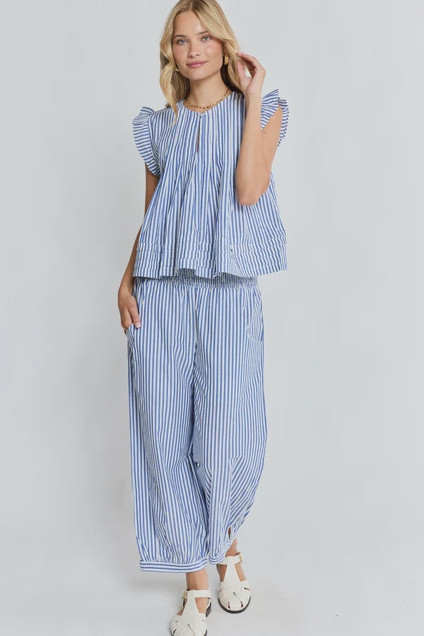 A woman modeling a matching blue and white striped two-piece set consisting of a ruffled short-sleeve top and wide-leg pants, paired with white sandals against a light gray studio background.