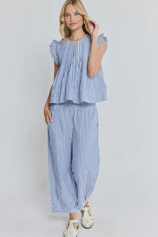A woman modeling a matching blue and white striped two-piece set consisting of a ruffled short-sleeve top and wide-leg pants, paired with white sandals against a light gray studio background.