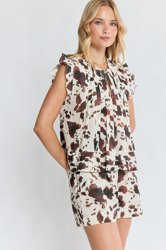 A woman modeling a sleeveless cream-colored dress with a brown and dark red cow-print pattern, featuring ruffled shoulders and a tie-neck detail, set against a plain light gray background.