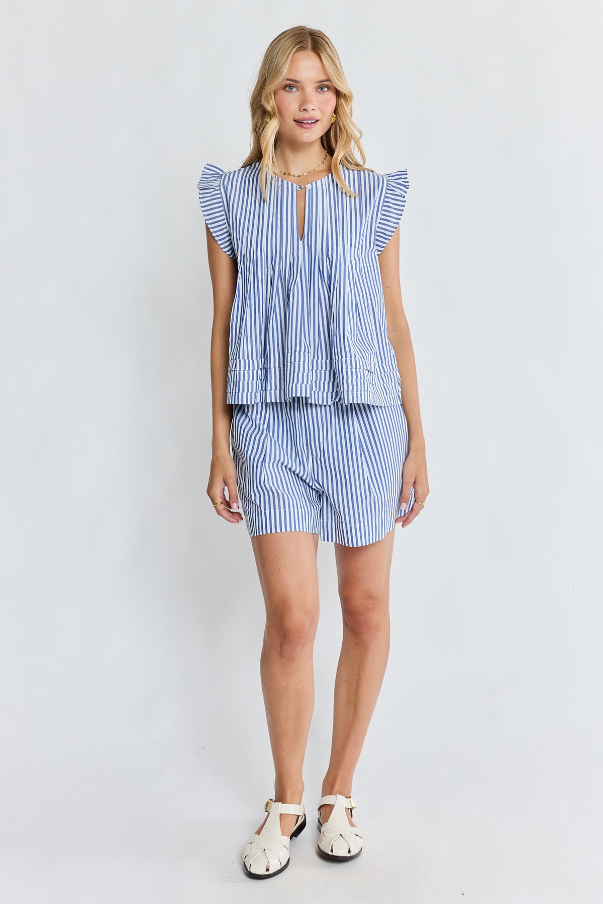 A woman modeling a matching blue and white striped two-piece set consisting of a ruffled short-sleeve top with a keyhole neckline and matching striped shorts, paired with white sandals, against a light gray studio background.