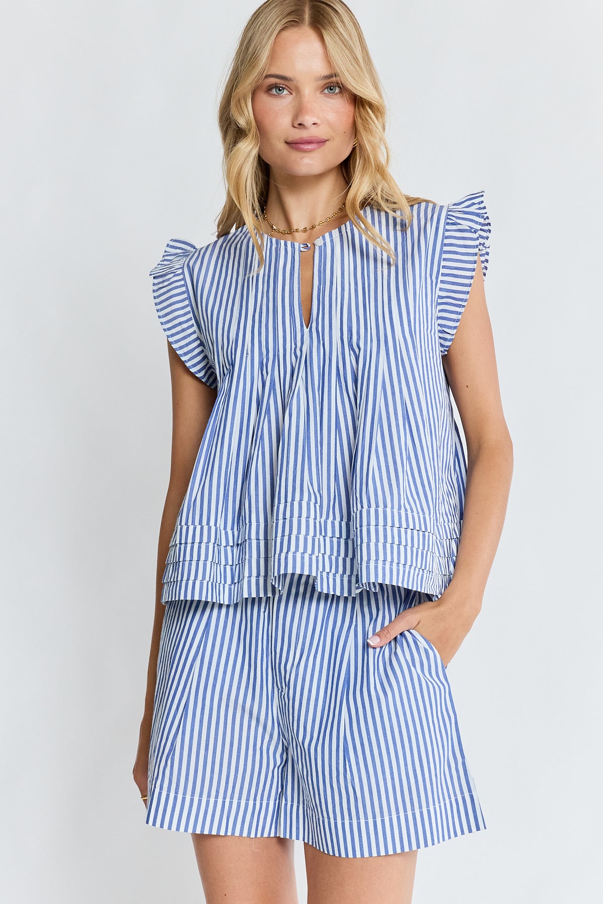 A woman modeling a matching blue and white striped two-piece set consisting of a ruffled sleeveless top with a keyhole neckline and pleated peplum hem, paired with matching striped shorts, against a light gray studio background.